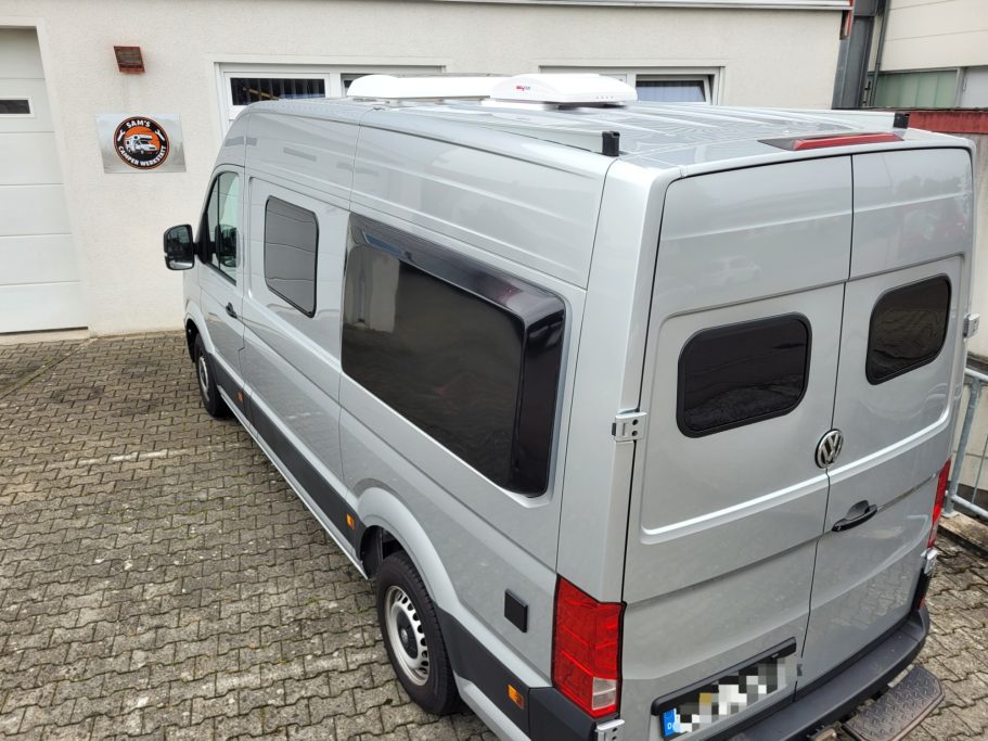 Silberner Lieferwagen mit Fenstern, hintere Ansicht auf einem gepflasterten Parkplatz.