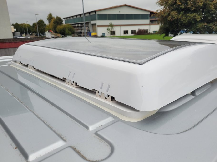Dachfenster mit Solarpanel auf einem Fahrzeugdach, im Hintergrund ein Gebäude.
