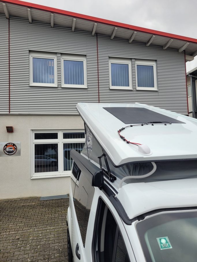 Eine Wohnmobilhaube mit Solarpanel vor einem grauen Gebäude mit Fenstern.