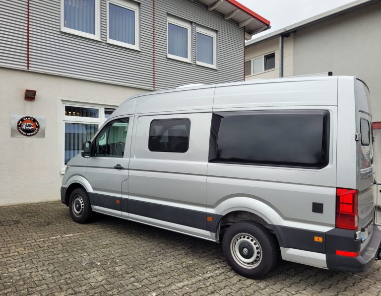 Fenstereinbau Camper Silberner Transporter parkt vor einem Gebäude mit grauer Fassade.