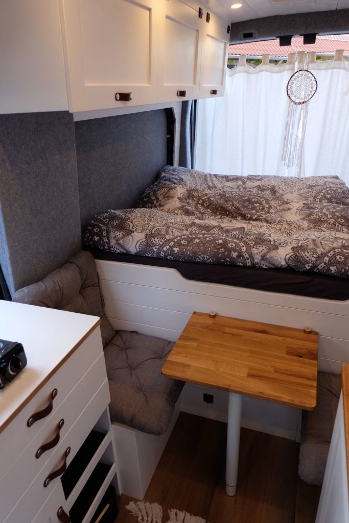 Kompakte Wohnmobileinrichtung mit Bett, Tisch und Stauraum in neutralen Farben.